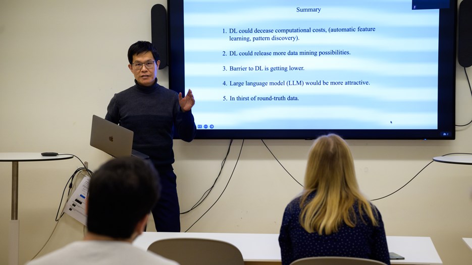 Jian-Feng Mao stands in front and to the side of a screen showcasing a summary slide. He is holding his laptop in one hand and gestures with the other.
