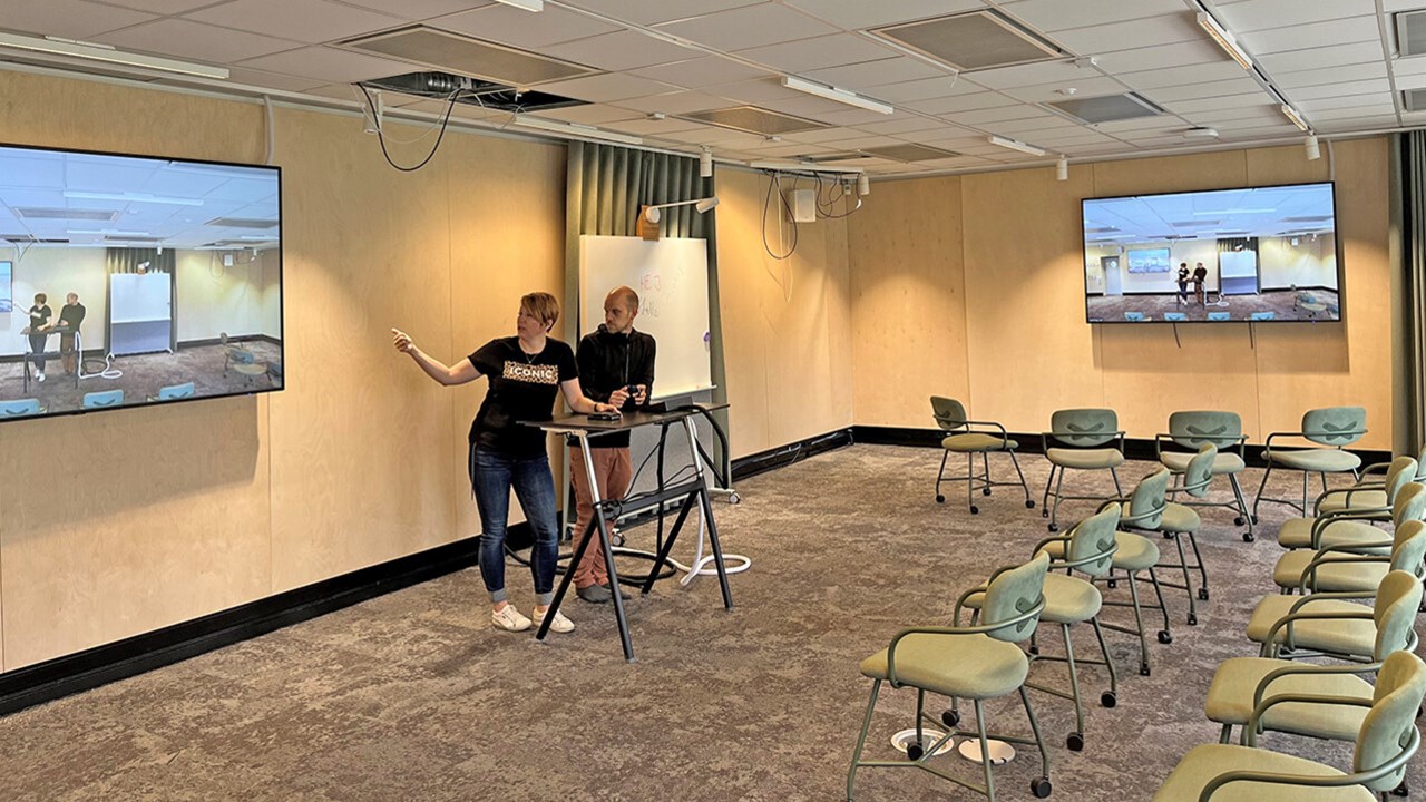 Åse Tieva och Jonas Lindholm, UPL, stand at a podium and look at themselves on a large screen to their left. On their right is a whiteboard and another large screen showing the same image.