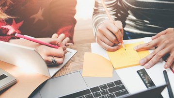 Två personer som sitter med datorer och block och skriver. Grupparbete.