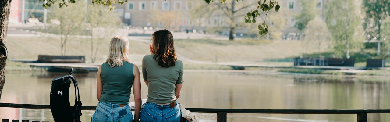 Två kvinnliga studenter sitter vid campusdammen. Ryggen emot kameran. Träd runtomkring.