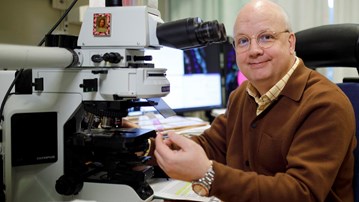 Thomas Brännström sitter vid sitt mikroskop och ler mot kameran med ett glas med vävnadssnitt i handen.