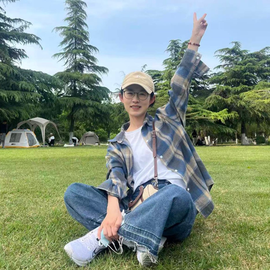 Xue-Chan Tian is sitting with crossed legs on a madow rising her right hand in the air showing a victory sign