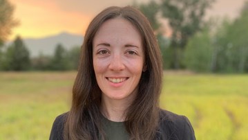A woman with long dark hair, wearing dark casually elegant clothes, isstanding in an open field during a colourful sunset.