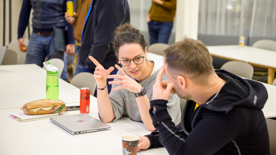 Barbora Parizkova speaks to a man with short hair and a black sweater. They are seated at a white table with a laptop, drinks and a sandwich in front of them. 