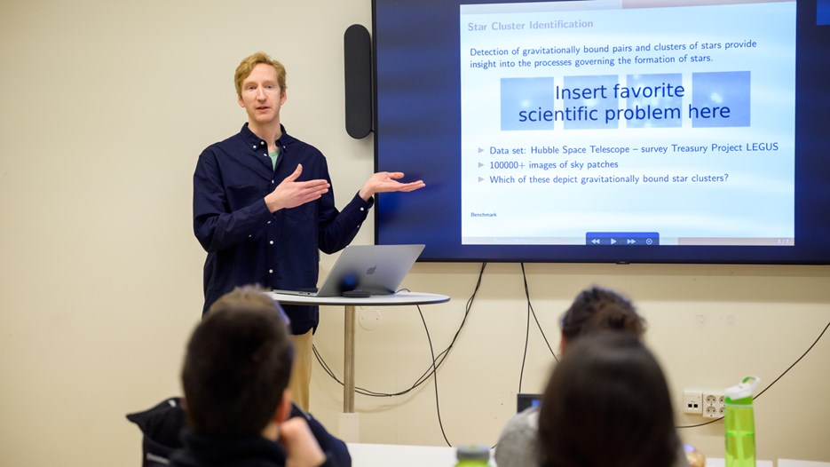 Jakob Hultgren speaks facing a seated crowd , to the side of him is a screen showing text: Insert favorite scientific problem here. 