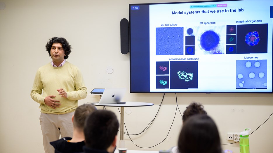 Aftab Nadeem speaks facing a seated crowd , to the side of him is a screen showing a slide with a human body and bacteria displayed.