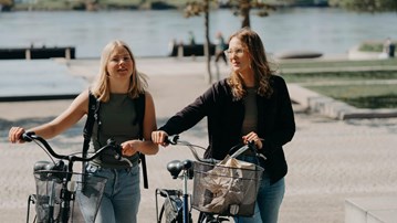 Två kvinnor går bredvid varandra och styr varsin cykel.