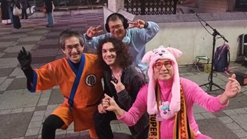 Gruppbild med tre personer som är utklädda i färgglada utstyrslar, som tillhör en teatertrupp, och en person som poserar tillsammans med dem.