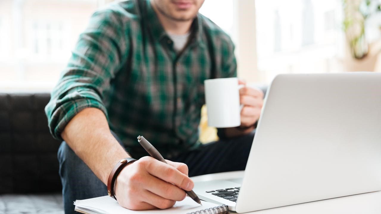 Person sitter i en soffa och arbetar. Har dator, kaffekopp och block där han skriver.