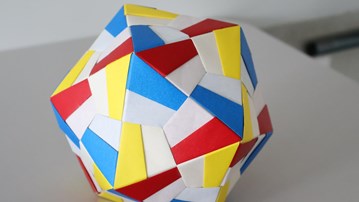 Färgglad modulär origami formad som en månghörnig boll, sammansatt av sammanflätade pappersbitar i rött, blått, gult och vitt, placerad på ett ljusgrått bord mot en neutral bakgrund.