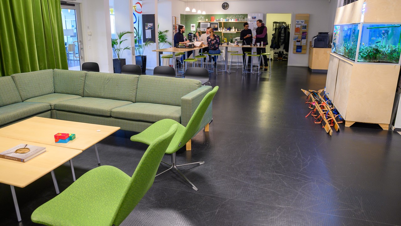 A living room and kitchen area  filled with furniture and a fish tank. A few people are gathered at the far end tables in the kitchen area.