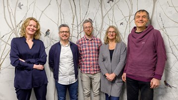 Five people standing in front of a wallpaper of a forest with birds