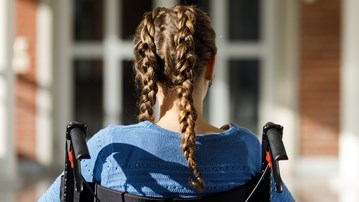 Flicka med flätor sitter i rullstol framför byggnad.