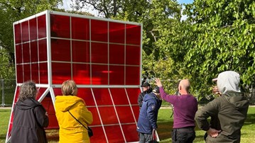 Människor står på en grön gräsmatta och beundrar en stor skulptur som liknar ett växthus i  rött glas.