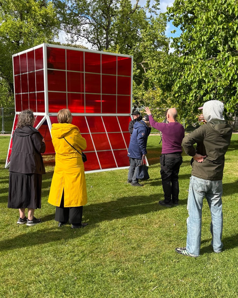 A group of people stand on green grass and marvel a big sculture that looks like a greenhouse in red glass.