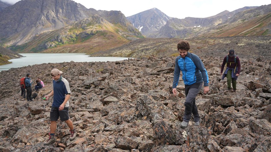 Three med hike on a stony path. In the background there is a lake and high mountains.