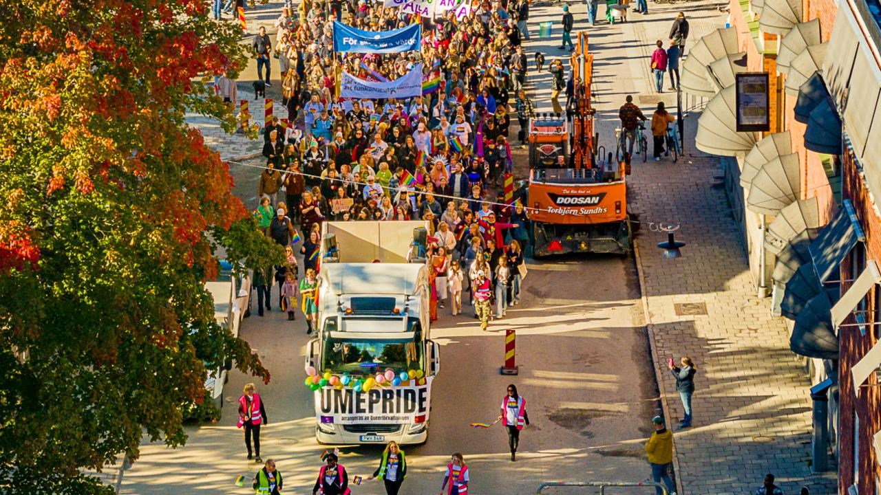 Dr&ouml;narbild av en parad av m&auml;nniskor som g&aring;r i Pride-t&aring;get i Ume&aring; 2024, med regnb&aring;gsflaggor och banderoller.