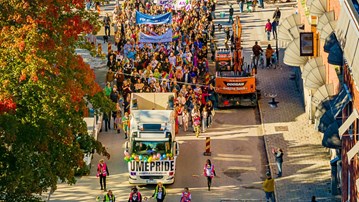 Drönarbild av en parad av människor som går i Pride-tåget i Umeå 2024, med regnbågsflaggor och banderoller.