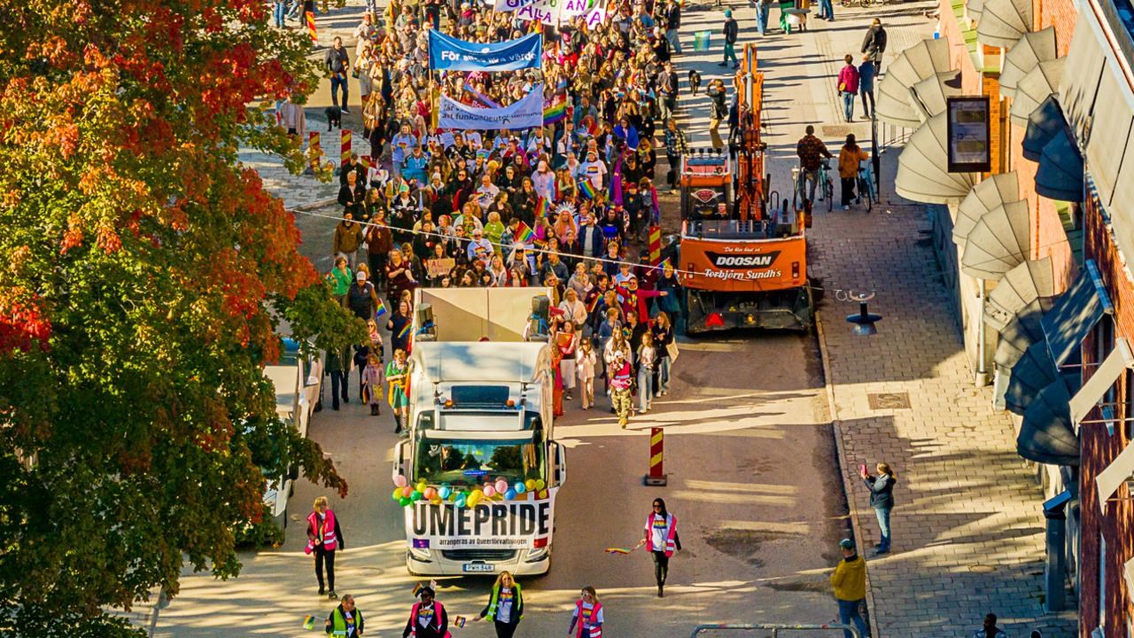Dr&ouml;narbild av en parad av m&auml;nniskor som g&aring;r i Pride-t&aring;get i Ume&aring; 2024, med regnb&aring;gsflaggor och banderoller.