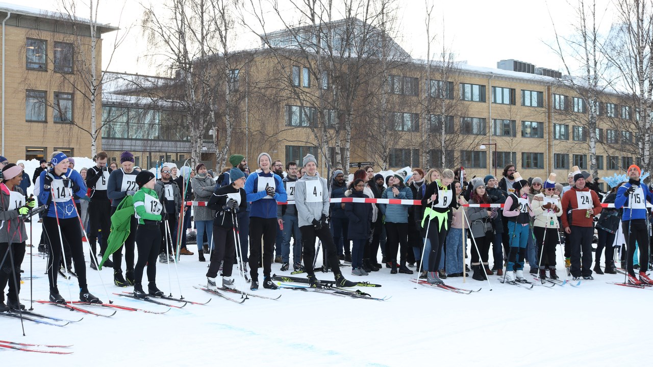 Several people on skis with numbers on their breast are waiting in front of  a white and red plastic band for their turn in the skiing relay. Behind of the plastic band, people cheering for the teams are standing.