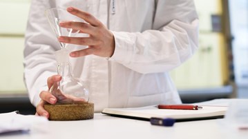 Detaljbild på student i labbrock som håller i glasbehållare.