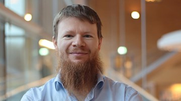 Alexandre Bartel i kort hår och skägg, står vid fönstret i ljusblå skjorta