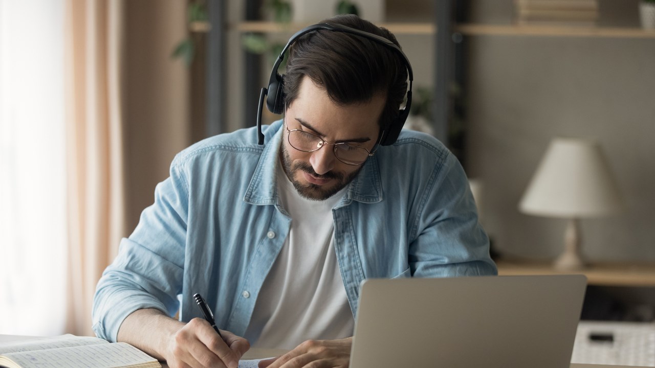 En man sitter vid ett bord med laptop och headset, böcker och antecknar i ett block.
