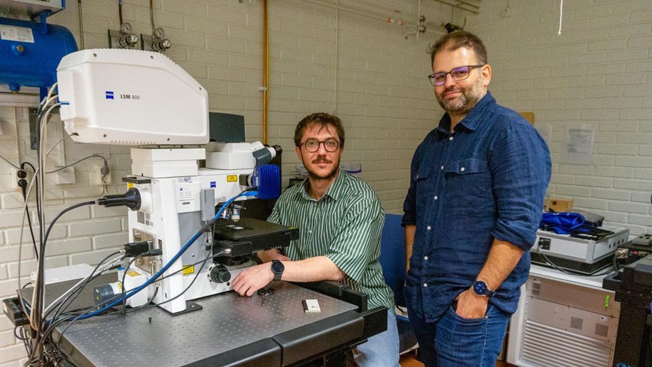 Two men at a huge microscope, one is sitting in front and one is standing next to him, both are looking into the camera.