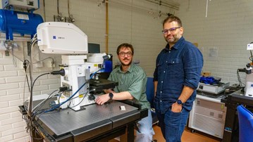 Two men at a huge microscope, one is sitting in front and one is standing next to him, both are looking into the camera
