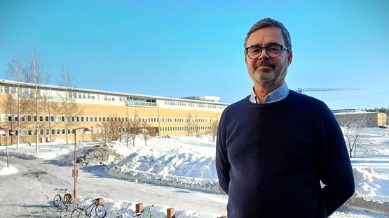 Man står utomhus med samhällsvetarhuset i bakgrunden. Solen lyser och det är vinter.