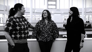 Tre kvinnor står och pratar och skrattar i en inomhusmiljö. Kortet är svartvitt.