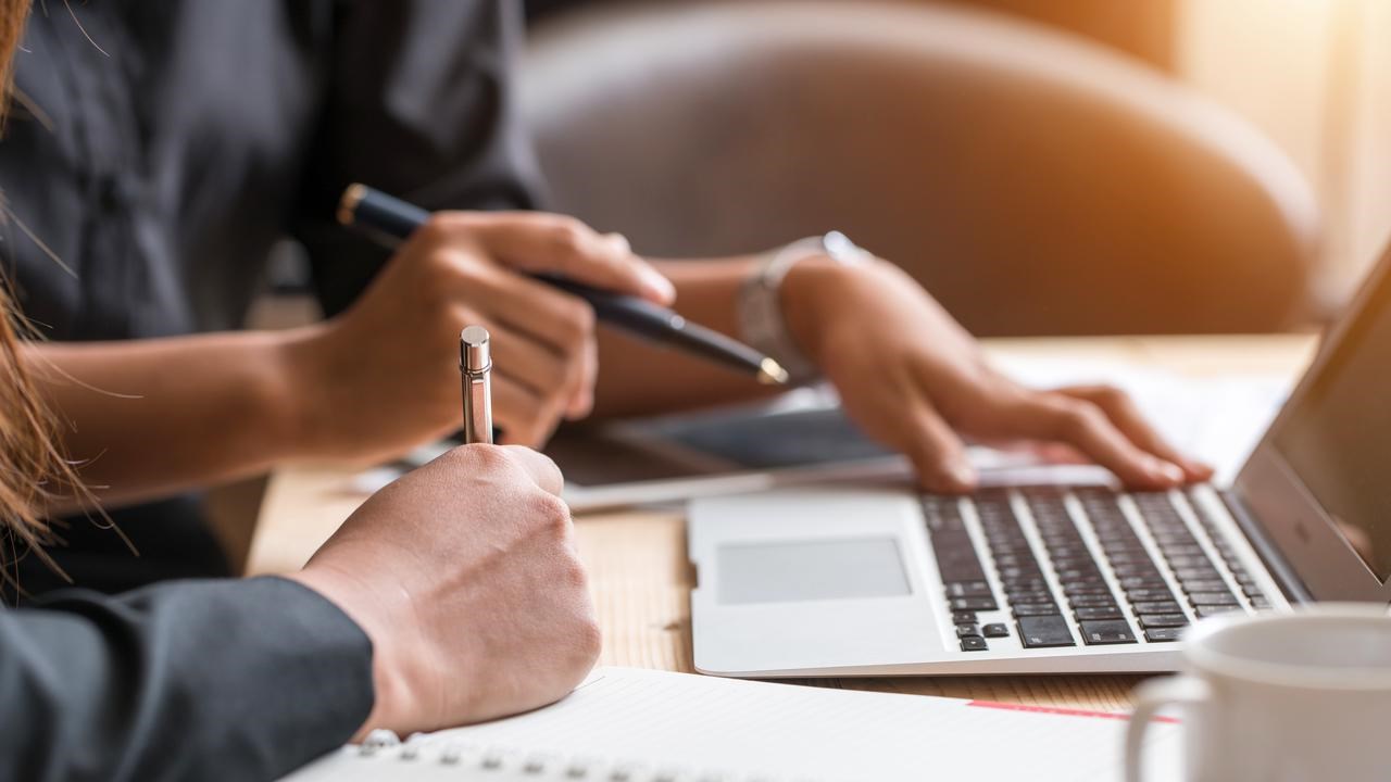 Närbild på två personers händer med dator, pennor och papper för att illustrera samarbete eller grupparbete.