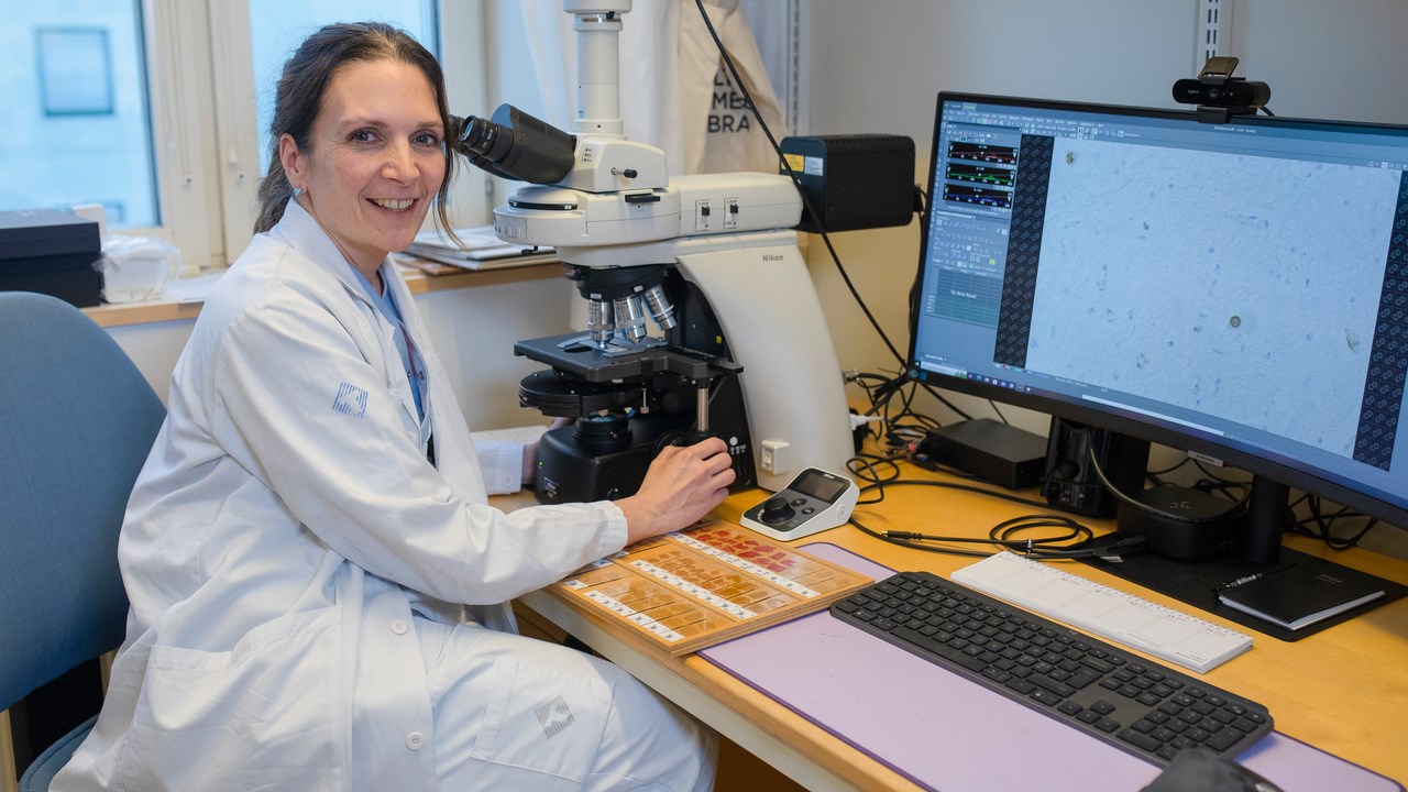 Karin Forsberg sitting at a microscope at her desk, facing the camera smiling. On her desk stained tissue samples mounted on glass slides are seen and on a computer screen a microscopy image of neuronal tissue is displayed.