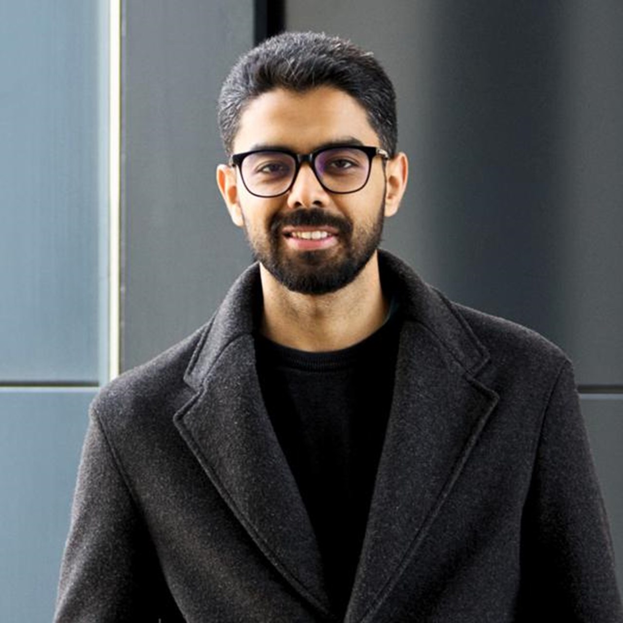 En man med sk&auml;gg och glas&ouml;gon st&aring;r framf&ouml;r en byggnad.