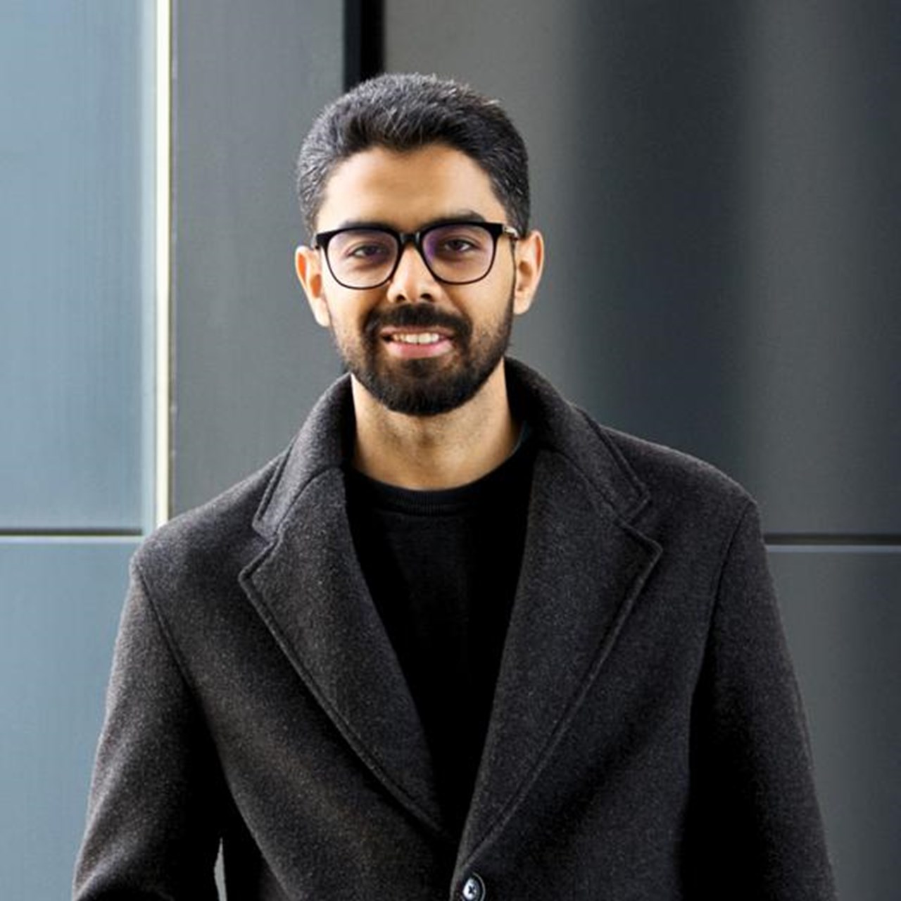 En man med sk&auml;gg och glas&ouml;gon st&aring;r framf&ouml;r en byggnad.