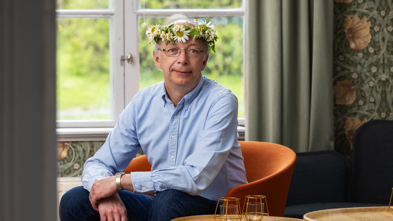Journalisten Arne Müller iklädd ljusblå skjorta, sitter i en fåtölj med en blomkrans runt huvudet.