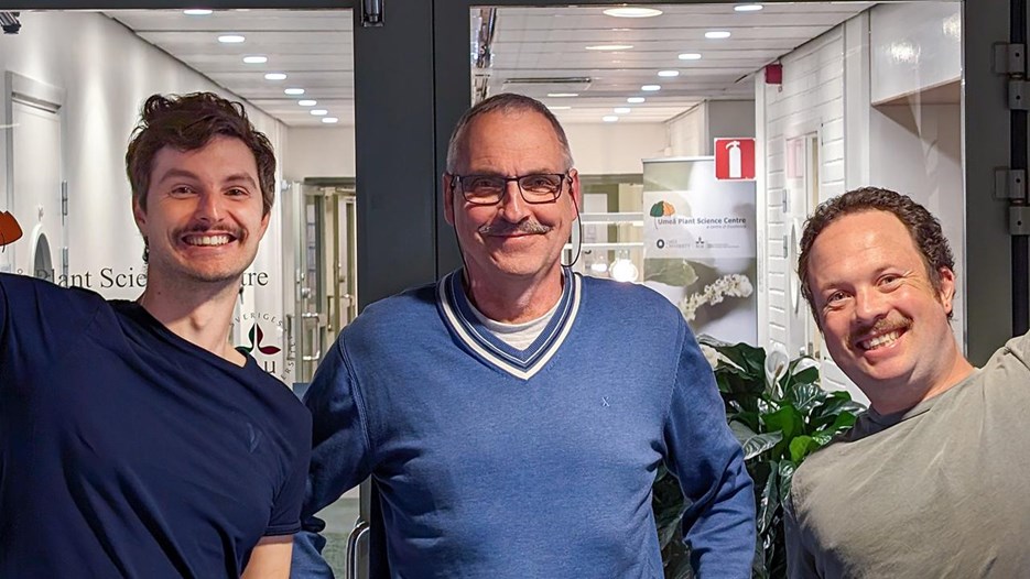 Tre män, alla med mustasch, ler in i kameran.