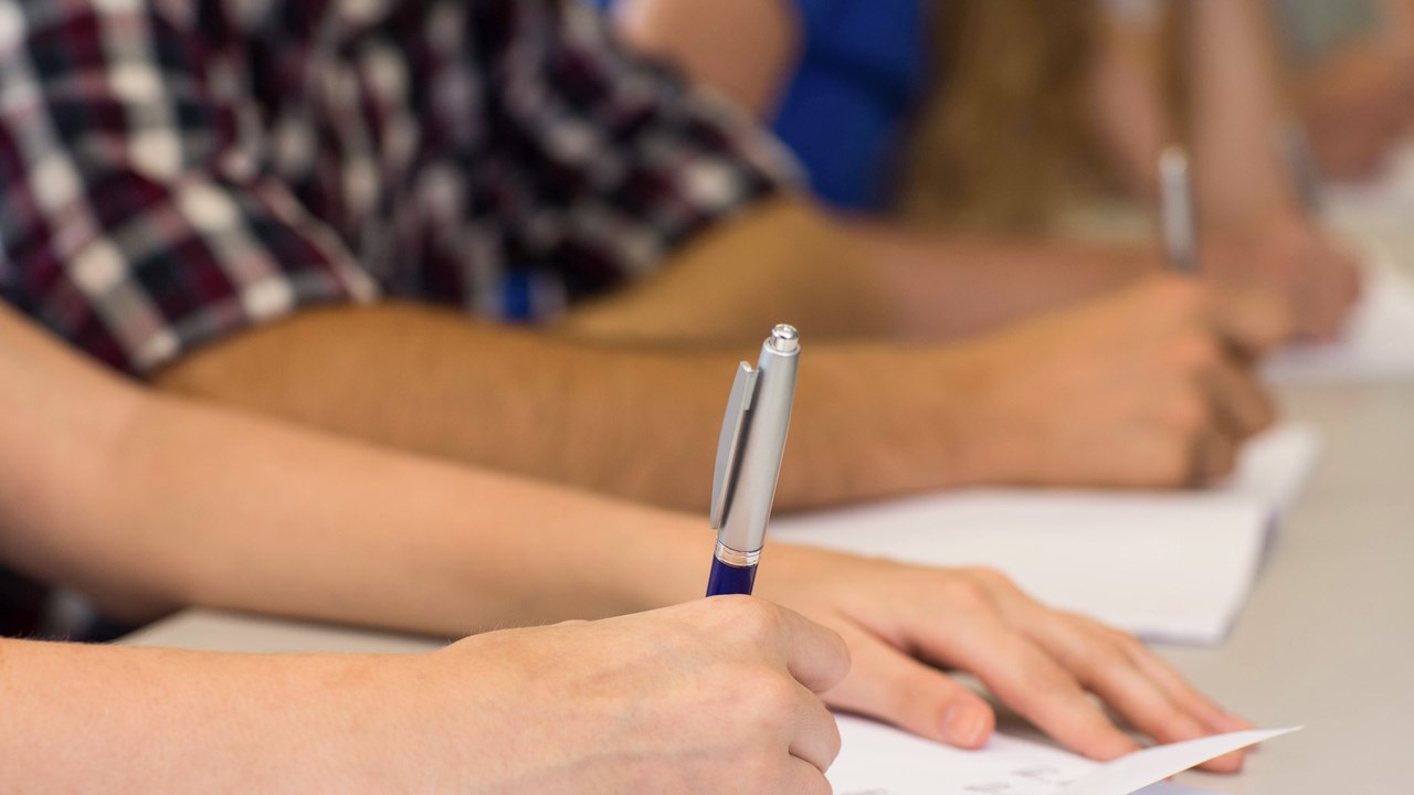 Detalj på händer som skriver på papper. Elever i klassrum.