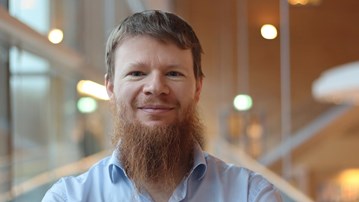 Alexandre Bartel i kort hår och skägg, står vid fönstret i ljusblå skjorta