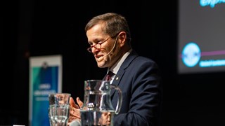 En man med kavaj och glasögon står vid ett podium och tittar ut mot publiken.
