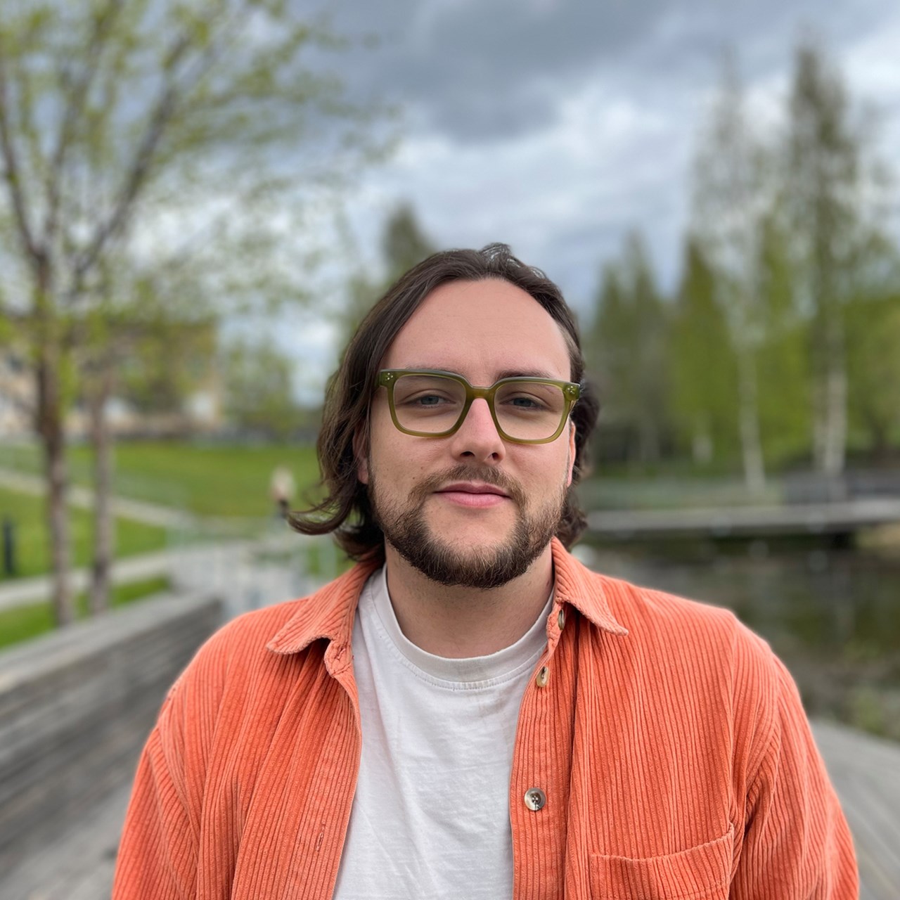 A man with long hair and glasses standing in front of a body of water.