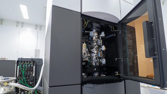 Elektronmikroskop som inhyses i en stor, 2x2x3 meter grå-vit behållare. Inuti syns mikroskopet med behållare och instrument av metall och många sladdar.