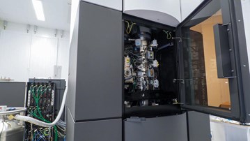 Elektronmikroskop som inhyses i en stor, 2x2x3 meter grå-vit behållare. Inuti syns mikroskopet med behållare och instrument av metall och många sladdar.