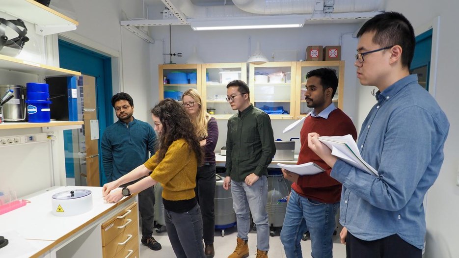 Students standing around the teacher in a lab, the teacher shows different tools to be used. Some students take notes. 