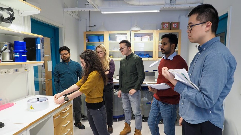 Students standing around the teacher in a lab, the teacher shows different tools to be used. Some students take notes. 