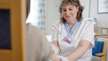 En person sitter med ryggen mot kameran och håller fram armen. Mitt emot sitter Elisabeth Müller Granberg i färd med att ta ett blodprov.