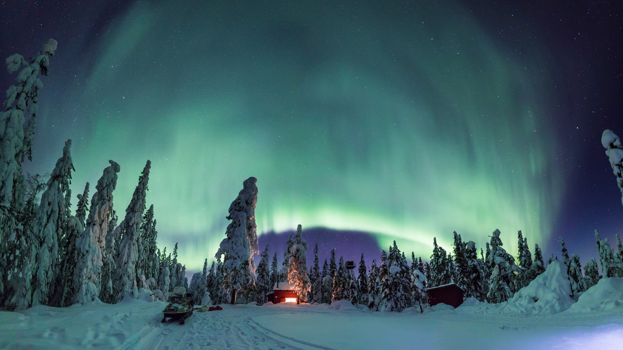 Upplyst stuga p&aring; vintern med ett stort gr&ouml;nt norrsken p&aring; den svarta himlen. Stugan st&aring;r vid skogsbryn.