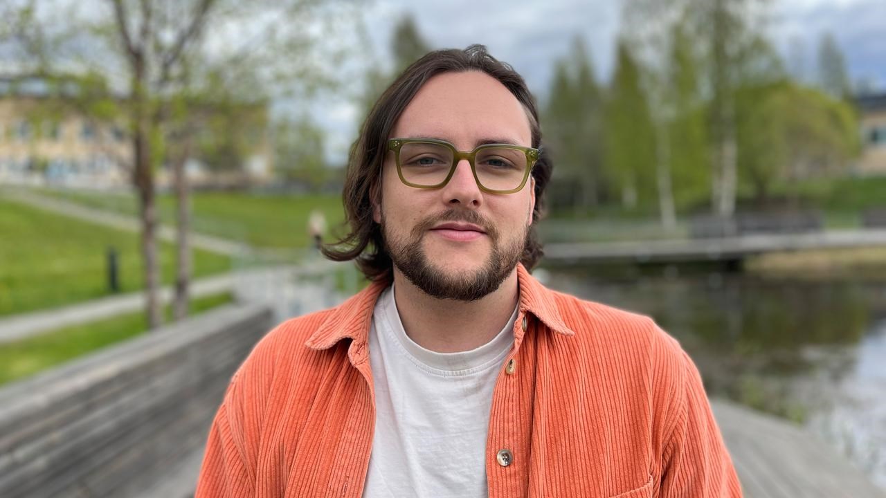 A man with long hair and glasses standing in front of a body of water.