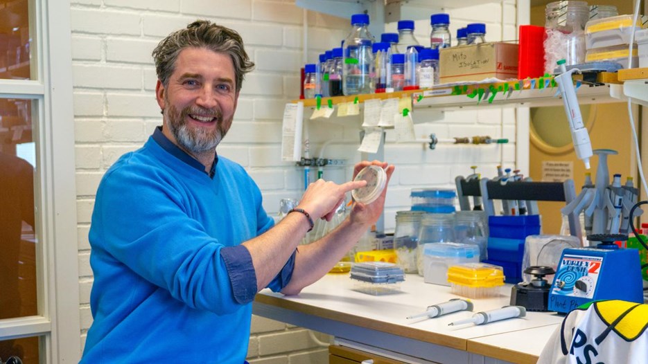 Olivier Keech wearing a blue sweater is sitting in a lab holding a round plate with blue spots in his one hand and is pointing with the small finger of his other hand on to it. He smiles into the camera.