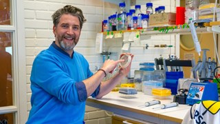 Olivier Keech wearing a blue sweater is sitting in a lab holding a round plate with blue spots in his one hand and is pointing with the small finger of his other hand on to it. He smiles into the camera.